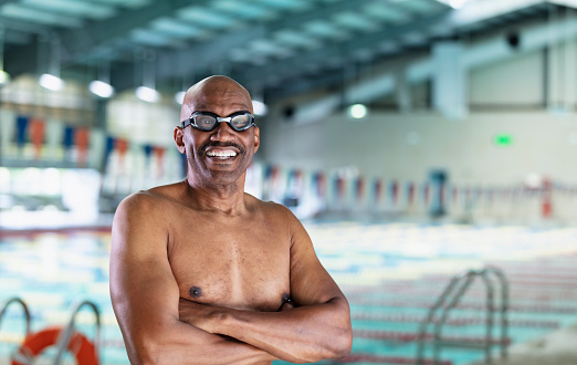 Headshot of a senior African-American man at an indoor swimming pool. He is standing on the deck looking at the camera, with the pool out of focus behind him. He is there to get exercise by swimming laps.