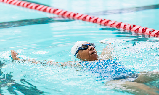 A senior African-American woman, in her 70s, swimming laps. She is in a lane, between two lane markers, swimming backstroke.