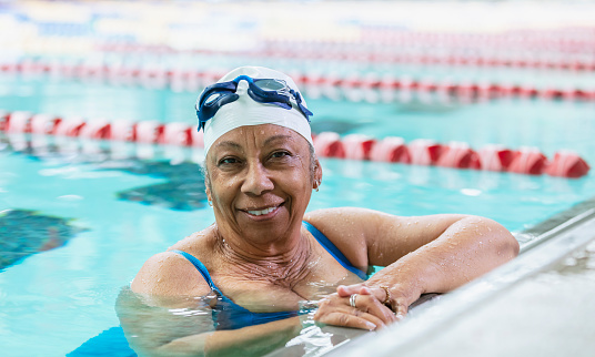 A senior African-American woman, in her 70s, swimming laps. She is the end of the lane, holding onto the wall, smiling at the camera after completing her workout.
