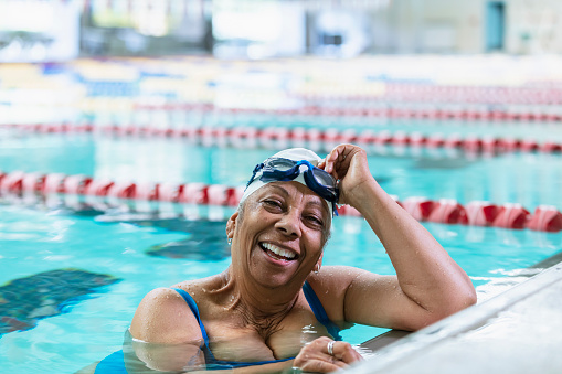 A senior African-American woman, in her 70s, swimming laps. She is the end of the lane, holding onto the wall, smiling at the camera and taking off her goggles after completing her workout.