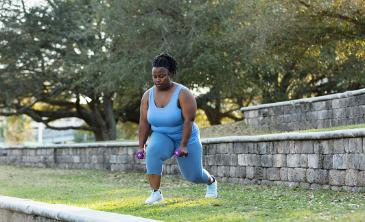 A mature African-American woman, in her 40s, exercising in the park. She is doing lunges. She is a plus size woman who looks fit and strong.