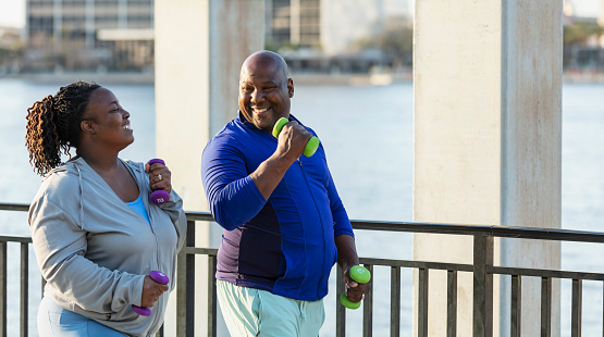 A mature African-American couple exercising together outdoors, power walking with hand weights on a city waterfront. They are smiling, enjoying their workout.