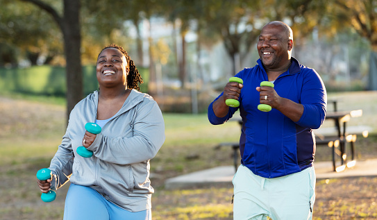 A mature African-American couple exercising in the park together, holding handweights. The woman is smiling at the camera and the man is looking at her.