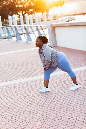 A mature African-American woman, in her 40s, exercising in the park. She is stretching and doing warm up exercises. She is a plus size woman who looks like she is in great shape, fit and strong.