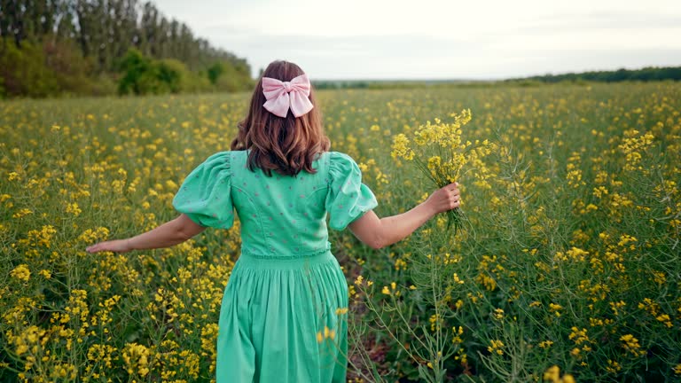 Back view of rural woman in turquose dress walks among blooming canola yellow flowers in field, amazing traveling footage. Romantic lady at springtime. Original lifestyle, floral background.