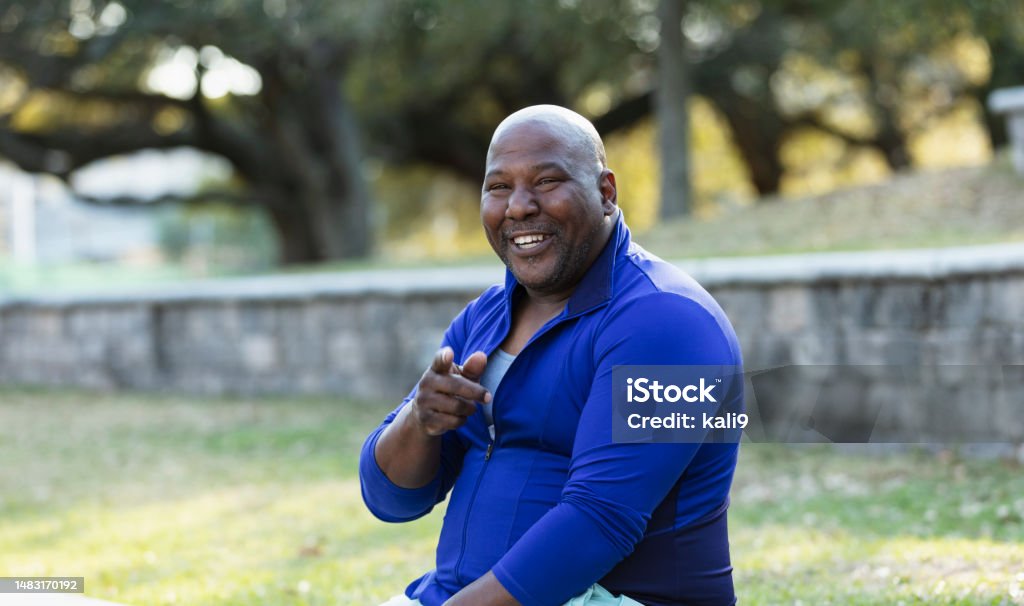 Mature African-American man at park, pointing at camera A mature African-American man sitting outdoors, at a park, on a sunny day, smiling and pointing his finger toward the camera. He is in his 40s with a shaved head and large build. Overweight Stock Photo