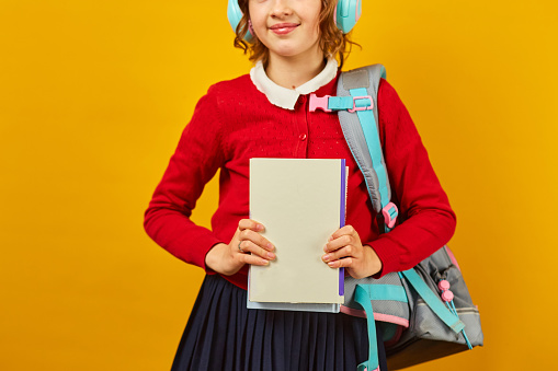 Unrecognizable Schoolgirl with backpack, headphone and books in hand back to school on yellow studio background, Teenager girl in school uniform.