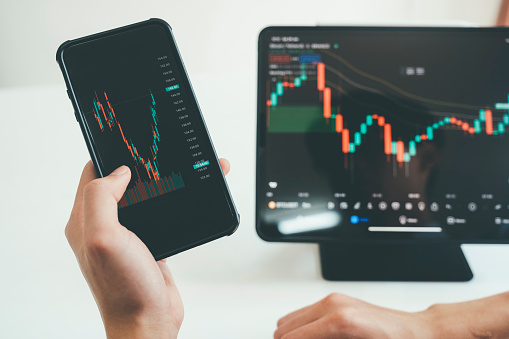Close-up of person's hands holding smartphone and tablet trading online on application, looking at screen, planning, and analyzing data to make decision. Online trading concept.