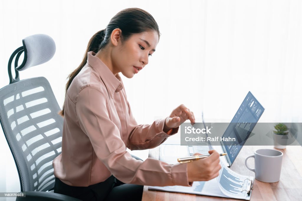 Young asian enthusiastic businesswoman at modern office desk with laptop. Young asian enthusiastic businesswoman at modern office desk using laptop to work and write notes. Diligent and attractive office lady working on computer notebook in her office work space. Adult Stock Photo