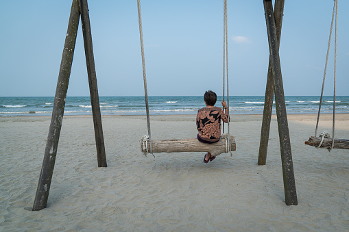 Back view of senior woman sitting on a wooden plank swing facing the sea. Retirement or travel vacation concept.