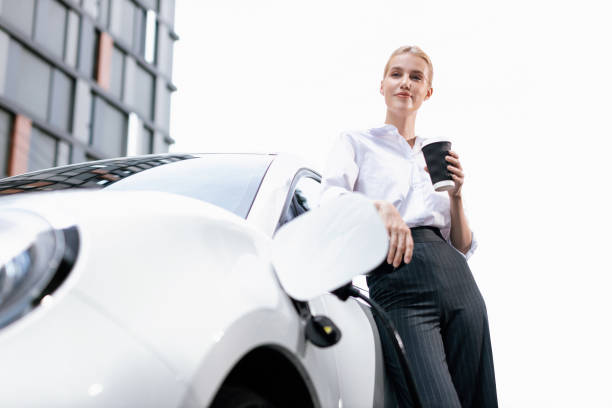 istock Progressive woman with coffee while charging EV car with residential buildings. 1483172313