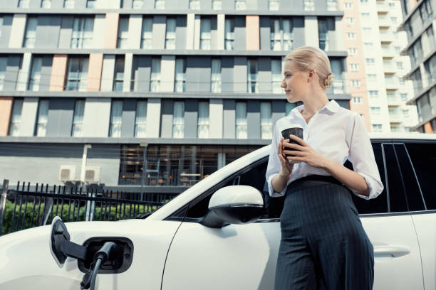 istock Progressive woman with coffee while charging EV car with residential buildings. 1483172347