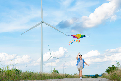 Progressive happy carefree boy concept. Young boy running along the road and flying kite at natural scenic on countryside, clear sky and sunny day with mountain and wind turbine background