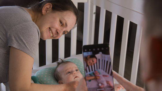 Father takes a picture or video of happy mother with newborn adorable daughter or son on mobile phone. A little baby in bodysuit lies in child crib. Concept of childhood, parenthood, love and family.