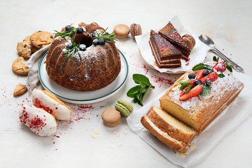 Dessert table with various of bundt cakes, pieces of cakes, sweets, cookies with fruit toppings on a white background. Top view.