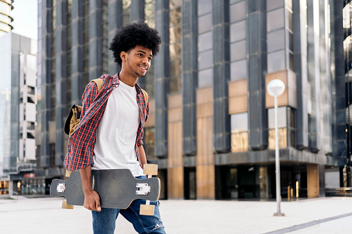Stock photo of young afro boy walking in the city carrying his longboard.
