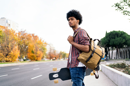 Stock photo of young afro boy walking in the city and carrying his longboard.