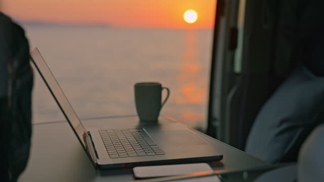 Interior of camper van nearby seascape during sunset. Laptop with mobile phone and coffee cup inside camper van parked in front of beautiful seascape during sunset.