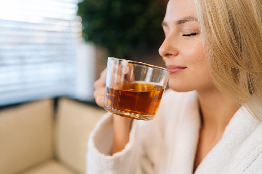 Close up face of charming young woman in white bathrobe drinking hot healthy tea with closed eyes after spa procedures. Relaxed blonde female resting with herbal drink sitting in couch at spa salon.