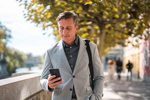 Waist up front view of a mature Caucasian businessman using a smart phone while walking outdoors in the city. He is wearing a formal suit and carrying a briefcase.