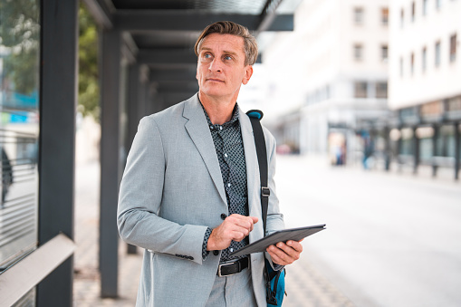 Good looking mature Caucasian businessman working on the go with a digital tablet. He is waiting at the bus station in the city, standing and looking away.