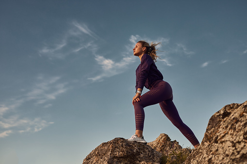 From below side view of fit female athlete stretching legs and warming up, before workout in nature while standing on stone against sunset sky and looking away
