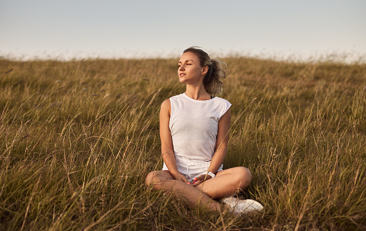 Young female in casual clothes closing eyes and breathing fresh air while sitting cross legged on grassy hill in windy evening