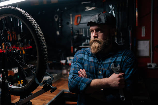 portrait d’un élégant réparateur barbu en casquette tenant une clé debout, les bras croisés à bicyclette, dans un atelier de réparation à l’intérieur sombre, regardant au loin. - cycling bicycle occupation men photos et images de collection