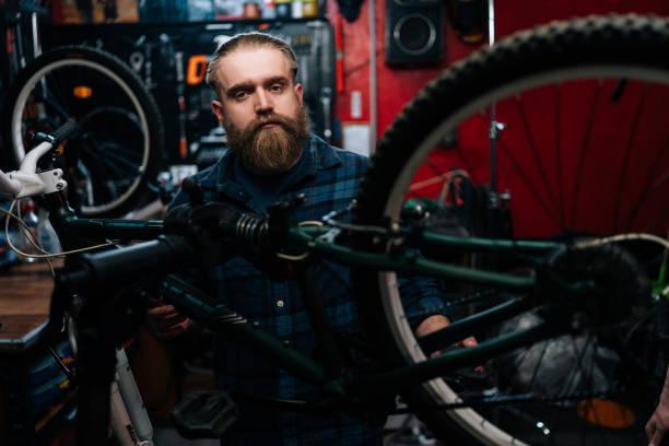 portrait d’un beau réparateur de vélos barbu avec une coupe de cheveux à la mode debout à vélo dans un atelier de réparation à l’intérieur sombre, regardant la caméra. - cycling bicycle occupation men photos et images de collection