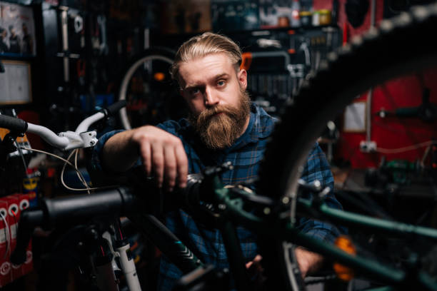 portrait d’un technicien barbu réparant un vélo de montagne debout sur un support à vélos travaillant dans un atelier de réparation de vélos avec un intérieur sombre, regardant la caméra - cycling bicycle occupation men photos et images de collection