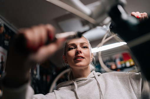 Closeup low-angle view of pretty blonde cycling mechanic female repairing and fixing mountain bicycle working in bike repair workshop with dark interior. Concept of professional maintenance of bicycle