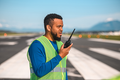 Portrait of a Hispanic Hispanic airfield operations  officer standing on a runway monitoring an airfield. Using a walkie talkie to communicate with an airplane and an air traffic control.