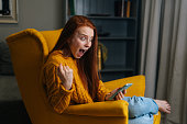 Portrait of happy excited young woman looking mobile phone screen sitting in yellow armchair, laughing with good news, feeling great positive surprise.