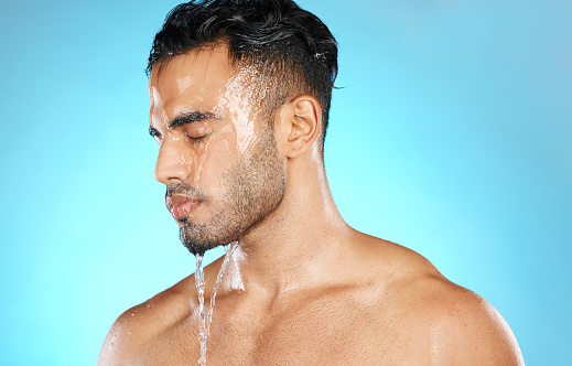 Shower, skincare and man in studio for water splash, hygiene and grooming on blue background. Cleaning, skin and Mexican model relax with luxury, moisture and wash routine while standing isolated