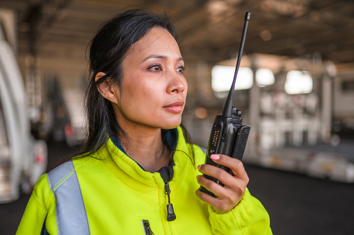 Portrait of an Asian female airport worker wearing a reflective vest and using a walkie-talki standing in a hangar coordinating.