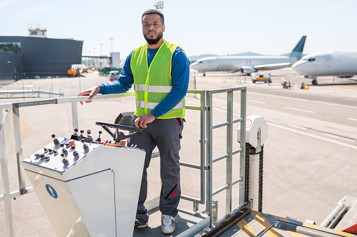 Hispanic male airport worker wearing a reflective vest driving and operating cargo loader on a runway.