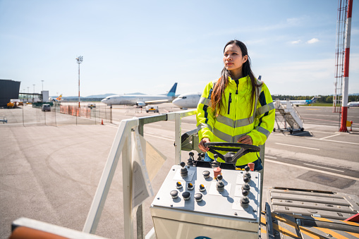 Asian female worker operating operating a container loader at the airport. She is wearing a reflective vest.