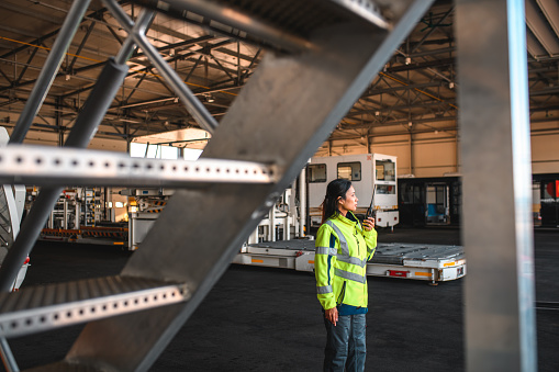 Asian female airport worker wearing a reflective vest and using a walkie-talki standing in a hangar coordinating.
