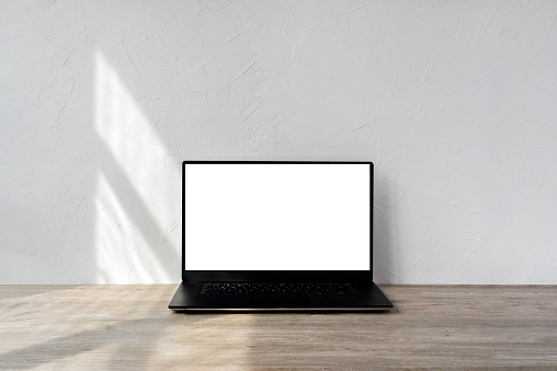 Laptop with blank screen mockup on a beige wooden table, geometric sunlight shadows on textured white wall, minimalist business, branding template, home interior design background