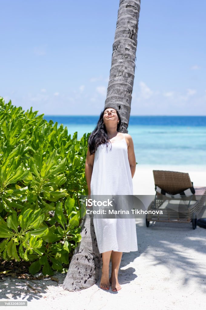 A young woman with long hair stands near a palm tree in Maldives and exposes her face to the sun A young woman with long dark hair stands near a palm tree in the Maldives and exposes her face to the sun. Vertical Stock Photo