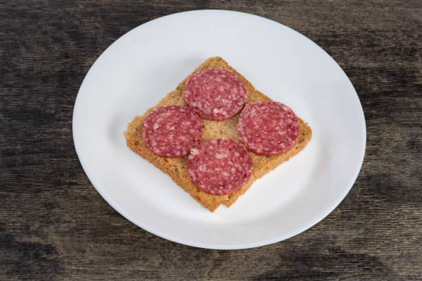Open sandwich made with multigrain bread and slices of the dry-cured sausage on white dish on a black surface
