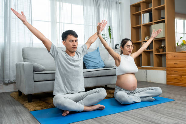 uomini e donne asiatici sono marito e moglie con mogli incinte che indossano camicie a ventre aperto. seduti in una posa yoga con le mani alzate sopra la testa per rilassarsi a piacimento nel mezzo del soggiorno. - torso physical therapy patient relaxation exercise foto e immagini stock