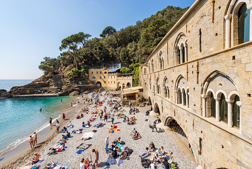 San Fruttuoso, Italy - April 8, 2023: Ancient San Fruttuoso Abbey (San Fruttuoso di Capodimonte), X-XI century, place of worship between Portofino and Camogli, Genoa province (Genova), Liguria, Italy, Europe. A large number of tourists sunbathe and relax on the small beach on a sunny spring day.