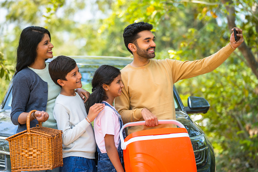 Happy smiling couple with kids taking selfie on mobile phone during summer camp picnic in front of car - concept of family trip, weekend holidays and togetherness