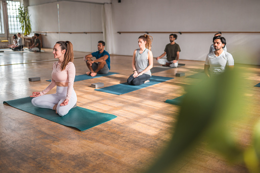 Candid shot of a group of various mid adult men and women sitting on yoga mats in lotus position and focusing on breathing at a yoga retreat. Full length image. Mirror in the background.