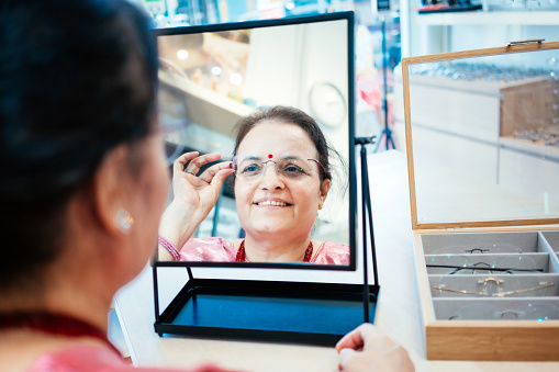 Female optician explaining to her senior woman customer trying on eyeglasses in front of mirror at optician store