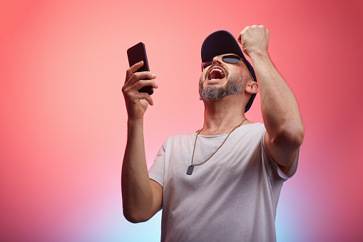 Man wearing casual clothing, t-shirt and a cap talking to phone super excited in front of colorful background