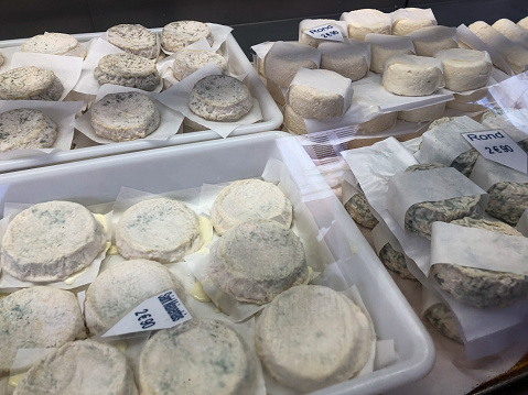 Horizontal high angle photo of white trays in a refrigerated window on a French food market stall, filled with a variety of round traditional cheeses. Saumur, Loire Valley, France.