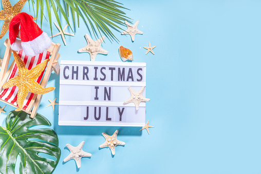 Christmas in July summer holiday or sale background. Summer vacation accessories, seashells, Starfish, lounge chair with Santa hat, tropical leaves. Getaway during winter holiday, top view copy space