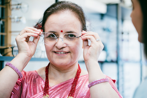 Female optician explaining to her senior woman customer trying on eyeglasses in front of mirror at optician store